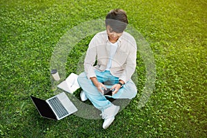 Asian man sitting on grass while using phone and laptop