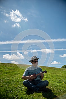 An Asian man sitting on a hill at a park, strumming a ukulele.
