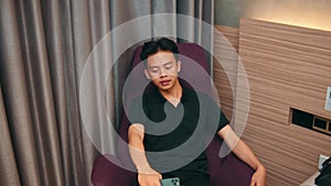 an Asian man is sitting on a purple sofa while waiting for his girlfriend in a hotel room