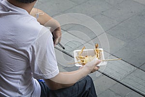 Asian man sitting in the street, holding a deep fried crab on a skewer, ready for eat