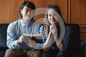 Asian man sitting teaching a Bible prayer to a Christian woman. As a confession of sins and blessing Jesus in background window.