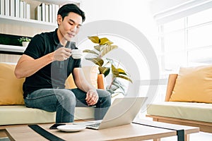 Asian man sitting and working on the sofa while drinking coffee. Businessman in casual working on laptop at home.
