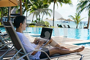 Asian man spent his summer vacation working on his laptop in a chair near the swimming pool in resort hotel near sea