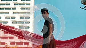 an Asian man standing in front of a building with a blue sky and red cloth flying