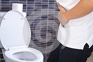 An Asian man stands near a toilet bowl. Have a stomachache, constipation in the bathroom. The handle on the stomach is sick due to