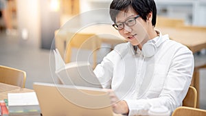 Asian man student reading book while using laptop in library