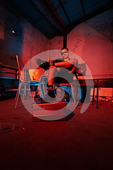 an Asian man with sunglasses sitting posing on a warehouse chair very sensually accompanied by a red light