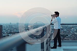 Asian man taking cityscape photo on building rooftop in low light situation. Photography, office people, or hobby concept