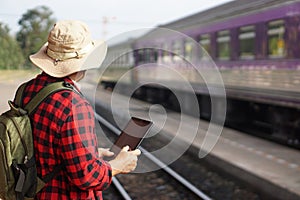 Asian man traveller is at railway station, wears hat, holds smart tablet.