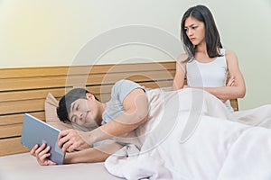 Asian man use taplet on bed, ignoring the girlfriend sitting next to him