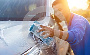 Asian man using blue sponge with soap to washing the car at outdoor in sunset time