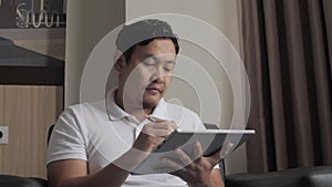 Asian man using pen stylus to draw on his digital tablet while sitting on couch in living room, young digital entrepreneur enjoys
