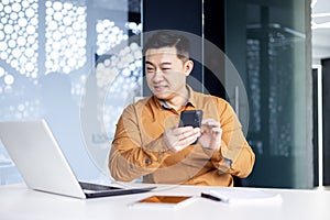 Asian man using phone in modern office, typing message and reading news while sitting at desk, businessman at work