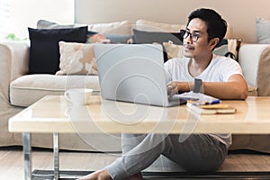 Asian man wearing glasses sitting on floor using laptop at home.