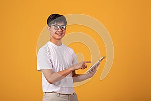 Asian man wearing glasses wearing a white shirt is standing with a tablet on orange background. Looking at camera