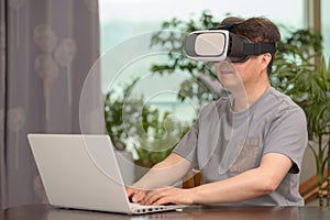 Asian man wearing VR headset and using laptop while sitting at his desk