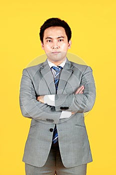 Asian man well dressed in a gray suit and white shirt, standing against a yellow background. A businessman Crossing his arms with