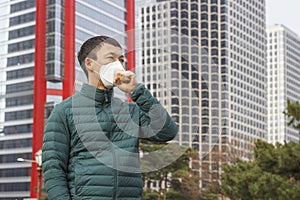 Asian man in white mask coughs and covers his face with his hand on the background of the city side view