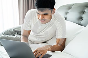 Asian man in white t-shirt laying on bed using laptop computer in bedroom