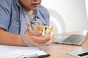 Asian man working and eating a burger on the office desk and heart attack. Concept of a busy businessman cannot work-left balance