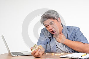 Asian man working and eating a burger on office desk and holding his neck after choking foods. Concept of a busy businessman