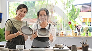 Asian mature woman and young woman holding ceramic pot from clay standing in pottery workshop