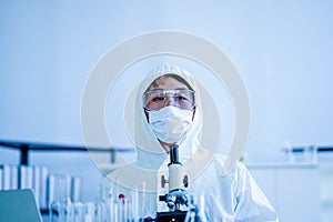 Asian medical scientist wearing protective white jumpsuit and face mask with glasses testing sample with microscope in clinical