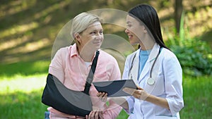 Asian medical worker with tablet smiling to injured senior patient in arm sling