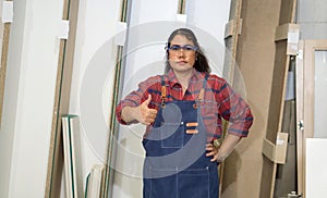 Asian middle aged woman with jean apron and protective glasses raise finger thumb up, stand infront plywood storage compartment.
