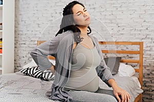 Asian mixed Caucasian pregnant woman suffering back pain sits on bed at home.