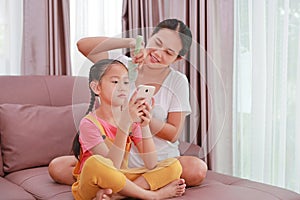 Asian mom combing hair for her daughter while playing smartphone on sofa in living room at home