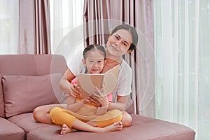 Asian mom and daughter reading a book on the sofa with looking at camera. Happy loving family at home