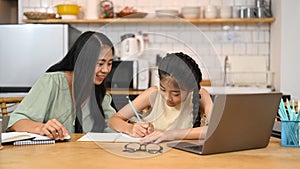 Asian mom helping her sweet daughter doing homework, learning online at virtual class on laptop computer at home