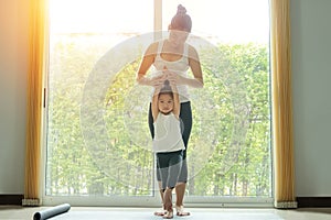 Asian mom practice yoga with a adorable daughter at home, try to teach young daughter to do yoga tree pose or vrksasana