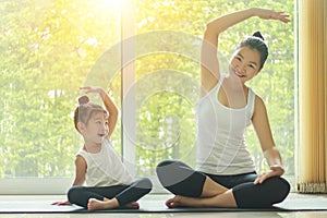 Asian mom practice yoga at home with a adorable daughter sitting next to her, trying to imitate the mother`s posture