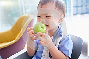 Asian 30 months / 2 years old toddler baby boy child sitting on high chair holding, biting, eating an unpeeled whole green apple a