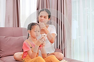 Asian mother braided hair for her little girl child while playing smartphone on sofa in living room at home