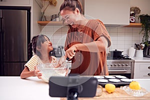 Asian Mother And Daughter Baking Cupcakes In Kitchen At Home Whilst On Vlogging On Mobile Phone photo