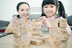 Asian mother and daughter playing wood block tower stacking game in cozy modern home. Ethnic babysitter engaging little girl in