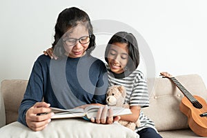 Asian mother and daughter read a book while sitting on the sofa. The little girl smiled and hugged his doll in the living room at