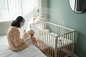 Asian mother embracing and playing with her baby son in bedroom. Young mom sitting on bed holding newborn child hands