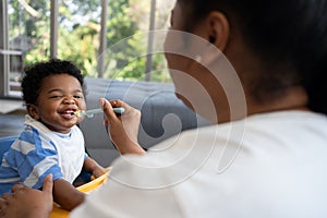 Asian mother feeding her 9 months old her cute little baby and African American helping for holding food plate At Home. Photo