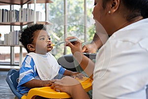 Asian mother feeding her 9 months old her cute little baby and African American helping for holding food plate At Home. Photo