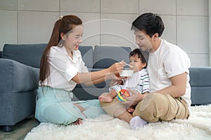 Asian mother feeding his 6 months old baby boy with solid food with spoon and father sitting near to cheer her son to eat food in