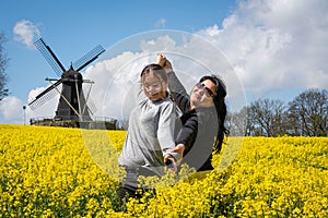 An asian mother in her 50s and her 11 years old daughter enjoys outing together in a field with yellow flowers and a windmill in t