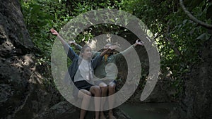 Asian mother and her teen daughter spread their arms happily with the wind blows while sitting on a huge rock in the forest.