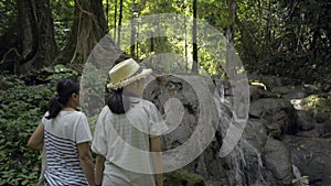 Asian mother and her teenage daughter wearing straw hat enjoy to see the beautiful scenery of small waterfall in tropical forest.