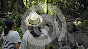 Asian mother and her teenage daughter wearing straw hat enjoy to see the beautiful scenery of small waterfall in tropical forest.