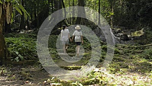 Asian mother and her teenage daughter wearing straw hat are walking hand in hand together in tropical forest with waterfall.