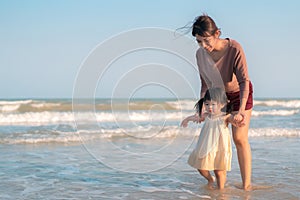 Asian mother holding hand daughter for support and walking on beach, Baby girl learning to walk, Happy family activity concept